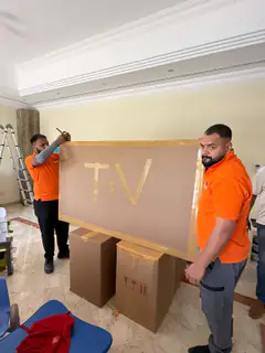 Two movers carrying a television in a labelled TV carton